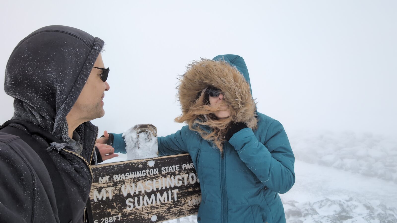 Two people at the Mt Washington Summit sign in blowing snow and whiteout conditions, hair and hood blasting sideways in the wind